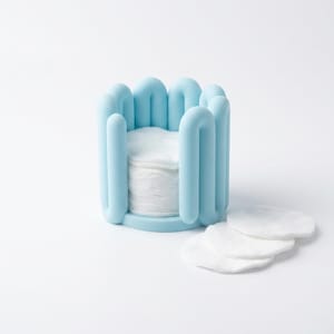 May include: A light blue cylindrical holder with a series of vertical, rounded loops forming the outer wall. Inside the holder is a stack of white, circular cotton pads, with three additional white cotton pads resting on the surface nearby.