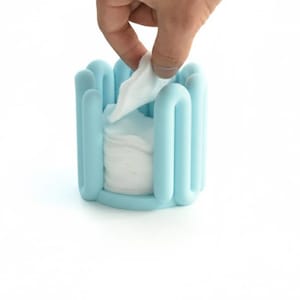 Puede incluir: Un dispensador de pañuelos de color azul claro con un diseño curvo único. El dispensador contiene pañuelos blancos, y una mano está sacando uno. El dispensador está hecho de un material suave y flexible.