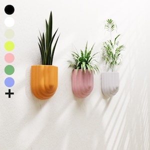 Op de afbeelding: Drie wandbevestigde plantenbakken met verschillende planten. De plantenbakken zijn gemaakt van een getextureerd materiaal en zijn oranje, roze en wit.