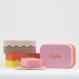 Op de afbeelding: Een verzameling kleurrijke zepen en zeepbakjes. De zepen zijn in tinten roze, oranje, geel en beige. Op een roze zeep staat de naam "Giulia". De zepen hebben een modern, minimalistisch design.