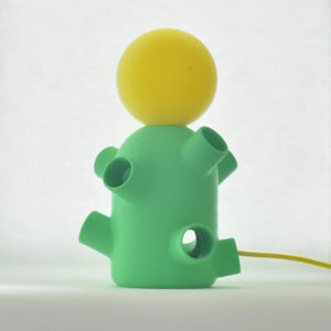 Könnte beinhalten: Eine grüne Tischlampe mit einer gelben Kugel oben. Die Lampe hat ein Kabel und mehrere Löcher im Sockel.