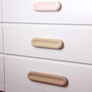以下が含まれることがあります： 3つの引き出しが付いた白いドレッサー。各引き出しには、それぞれ異なる色の細長い取っ手が付いています。一番上の取っ手は淡いピンク、真ん中はベージュ、一番下は茶色です。取っ手は丸みを帯びていて滑らかです。