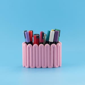 Peut inclure: Un organisateur de bureau rose clair avec une structure murale ondulée et serpentine continue. Le récipient est rempli d'une variété de marqueurs et de stylos de couleurs assorties, notamment rouge, violet, blanc, vert et noir, tous verticaux.