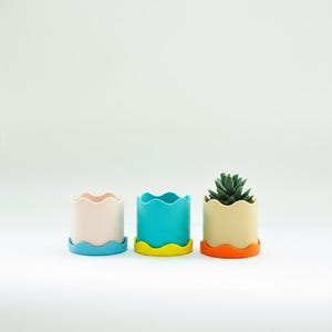 Peut inclure: Trois petits pots de fleurs colorés avec des bords ondulés et des bases assorties. Les pots sont dans des tons pastel de rose, de turquoise et de beige, avec des bases bleues, jaunes et oranges. Un pot contient une petite plante succulente.