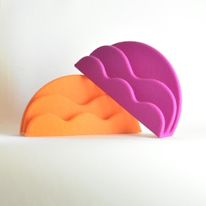 Op de afbeelding: Twee halfronde, golfvormige decoratieve objecten. De ene is feloranje, de andere magenta. De objecten zijn gemaakt van een zacht, viltachtig materiaal en staan tegen een witte achtergrond. Het ontwerp is modern en abstract.