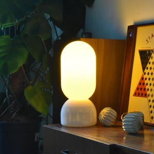 Op de afbeelding: Een witte keramische tafellamp met een ronde, matte glazen kap. De lamp is aan en geeft een warm licht. De lamp staat op een houten tafel naast een spelbord.