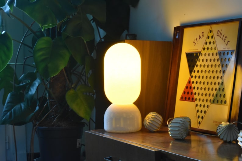 Puede incluir: Una l&aacute;mpara de mesa de cer&aacute;mica blanca con una pantalla redonda de vidrio esmerilado. La l&aacute;mpara est&aacute; encendida y proyecta un brillo c&aacute;lido. La l&aacute;mpara est&aacute; sobre una mesa de madera junto a un tablero de juego.