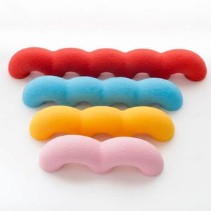 Puede incluir: Un conjunto de cuatro objetos ondulados y texturizados dispuestos en una pila. La pieza superior es roja, seguida de una pieza azul, una amarilla y una rosa en la parte inferior. Cada objeto presenta una forma curva repetitiva y mate.