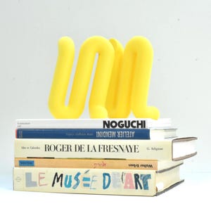 Può includere: Una scultura astratta giallo brillante a forma di lettere "UM" è posta sopra una pila di libri. I libri hanno titoli come "Noguchi" e "Le Musée D'Art". La scultura aggiunge un tocco di colore allo sfondo neutro.