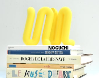 Puede incluir: Una escultura abstracta amarilla brillante con forma de las letras "UM" se encuentra sobre una pila de libros. Los libros tienen títulos como "Noguchi" y "Le Musée D'Art". La escultura añade un toque de color al fondo neutro.