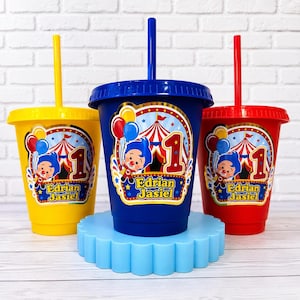Puede incluir: Tres vasos de plástico con tapas y pajitas en amarillo, azul y rojo. Cada vaso tiene una pegatina circular con una carpa de circo, un personaje de dibujos animados, el número 1 y el texto Edrian Jasiel en letras amarillas y azules.