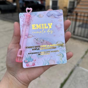 Puede incluir: Un frasco de burbujas rosa con una parte superior en forma de corazón está al lado de una tarjeta con el texto "EMILY cumple dos" en oro. La tarjeta tiene un tema submarino con ilustraciones de ballenas, medusas e hipocampos. La tarjeta también tiene texto en español.