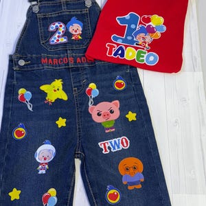 Puede incluir: Un conjunto de peto vaquero y camiseta roja. El peto tiene parches de personajes de dibujos animados, el número "2" y el texto "MARCOS AD". La camiseta roja presenta el número "1" y el nombre "TADEO".