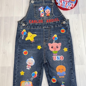 Puede incluir: Peto vaquero azul oscuro decorado con personajes de dibujos animados, globos y estrellas. El peto tiene el nombre "CARLOS ABDIEL" en letras rojas y el número "1". También se incluye un gorro rojo y azul a juego con un pompón rojo.