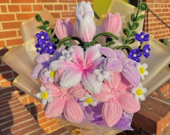 May include: A bouquet of pink, purple and white felt flowers wrapped in cream-coloured paper with a gold ribbon. The bouquet is held by a hand wearing a beige jacket.