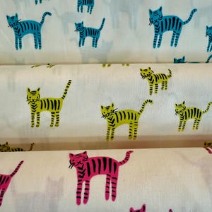 May include: White fabric rolls with a repeating pattern of stylized cats in blue, yellow, and pink. The cats have striped bodies and are arranged in a playful, repeating design. The fabric appears to be cotton or a similar material.