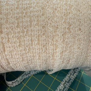 May include: A roll of cream-colored lace fabric with a delicate floral pattern. A length of the same lace is unrolled, showing the intricate design. The fabric is soft and textured, suitable for various craft projects.