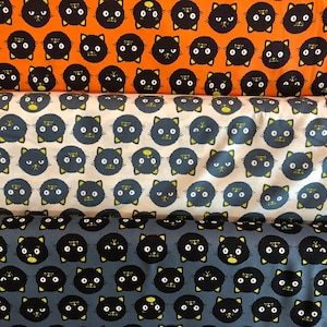 May include: Three fabric swatches with a repeating pattern of black cat faces. The top swatch is orange, the middle swatch is cream, and the bottom swatch is blue.