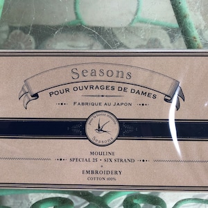 May include: A brown cardboard box with a black border and white text that reads "Seasons Pour Ouvrages de Dames Fabrique au Japon" and "Mouline Special 25 Six Strand Embroidery Cotton 100%". A small blue and white logo with a bird is in the center of the box.