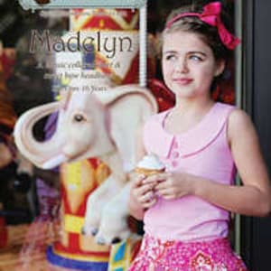 May include: A young girl wearing a pink sleeveless shirt with a white collar and a pink and white patterned skirt. She is holding a cupcake and smiling at the camera. The image is set in front of a carousel with a white elephant.