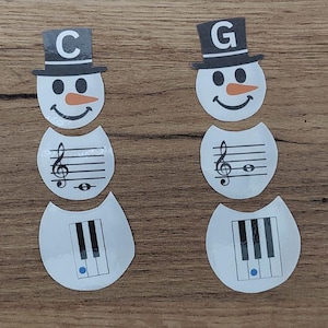 Op de afbeelding: Drie sneeuwmanvormige papieren uitsparingen met muzikale thema's. Elke sneeuwman heeft een zwarte hoge hoed met een letter, een wortelneus en een lachend gezicht. De lichamen bevatten muzieknoten en pianotoetsen.