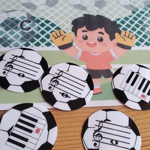 May include: A colorful educational game featuring soccer ball-shaped cards with musical notes and piano keys. A cartoon goalkeeper stands in front of a soccer net. The background includes soccer ball graphics.