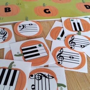 May include: A collection of square cards featuring orange pumpkin illustrations with black and white musical notation, including treble clefs, bass clefs, and piano keys, alongside a larger rectangular sheet with pumpkins labeled with letters.