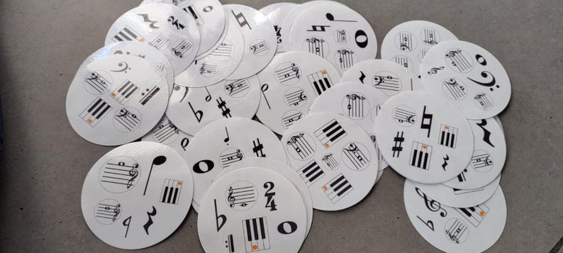 May include: A collection of white, circular music-themed stickers scattered on a gray surface. Each sticker features black musical notes, clef symbols, and piano keys. Some stickers have orange accents.
