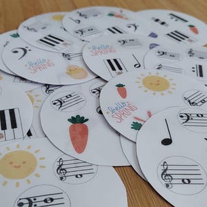 May include: A collection of white, circular stickers with spring-themed illustrations. Designs include musical notes, piano keys, carrots, chicks, and smiling suns. Some stickers have the text "Hello Spring".