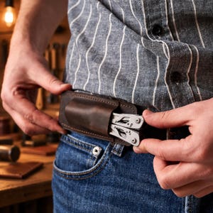 Puede incluir: Una funda de cinturón de cuero marrón oscuro que contiene una herramienta multifunción de acero inoxidable plateado. La funda está fabricada con costuras visibles y se sujeta a un cinturón sobre unos vaqueros azules y una camisa a rayas.