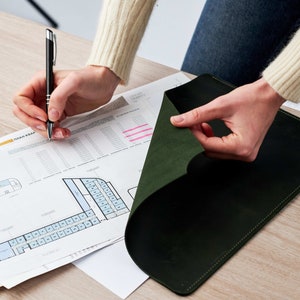 May include: A person is reviewing a green leather portfolio folder. The folder is open and the person is using a pen to mark a document with a floor plan labeled "План Квар", which is Russian for "Floor Plan".