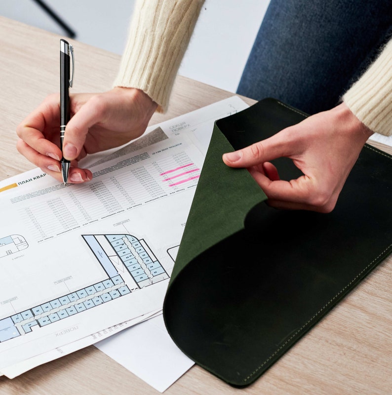 May include: A person is reviewing a green leather portfolio folder. The folder is open and the person is using a pen to mark a document with a floor plan labeled "План Квар", which is Russian for "Floor Plan".