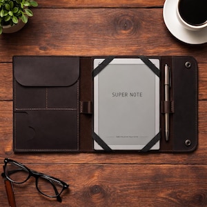 May include: A dark brown leather folio case with a Super Note tablet, pen, and elastic straps. The tablet is grey with the words "SUPER NOTE" printed on the screen. The folio is open on a wooden surface with a cup of coffee and glasses.