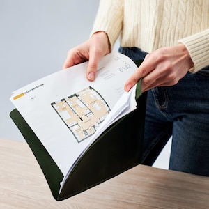 May include: A person is holding a green folder with a white sheet of paper inside. The paper has a floor plan of an apartment with the text "План квартиры" in yellow at the top.