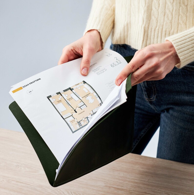 May include: A person is holding a green folder with a white sheet of paper inside. The paper has a floor plan of an apartment with the text "План квартиры" in yellow at the top.