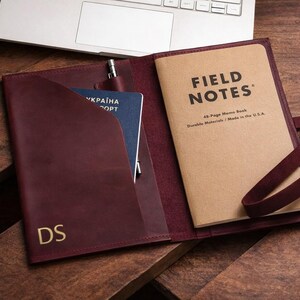 May include: Burgundy leather notebook cover with a Field Notes memo book and a passport. The cover has a pen holder and a strap. The initials "DS" are embossed in gold on the front. A laptop is visible in the background.
