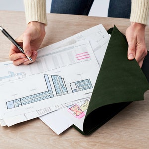May include: A person is reviewing a set of architectural plans, including a floor plan with a blue and white color scheme. The plans are laid out on a wooden table and are being held down by a dark green folder.