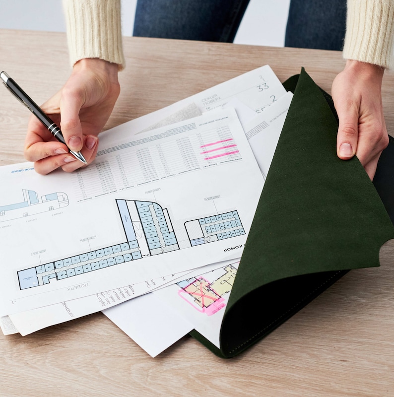 May include: A person is reviewing a set of architectural plans, including a floor plan with a blue and white color scheme. The plans are laid out on a wooden table and are being held down by a dark green folder.