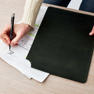 May include: A person signing a document with a black pen. The document is a blueprint with a green highlighter marking a section. The person is sitting at a desk with a black leather portfolio.