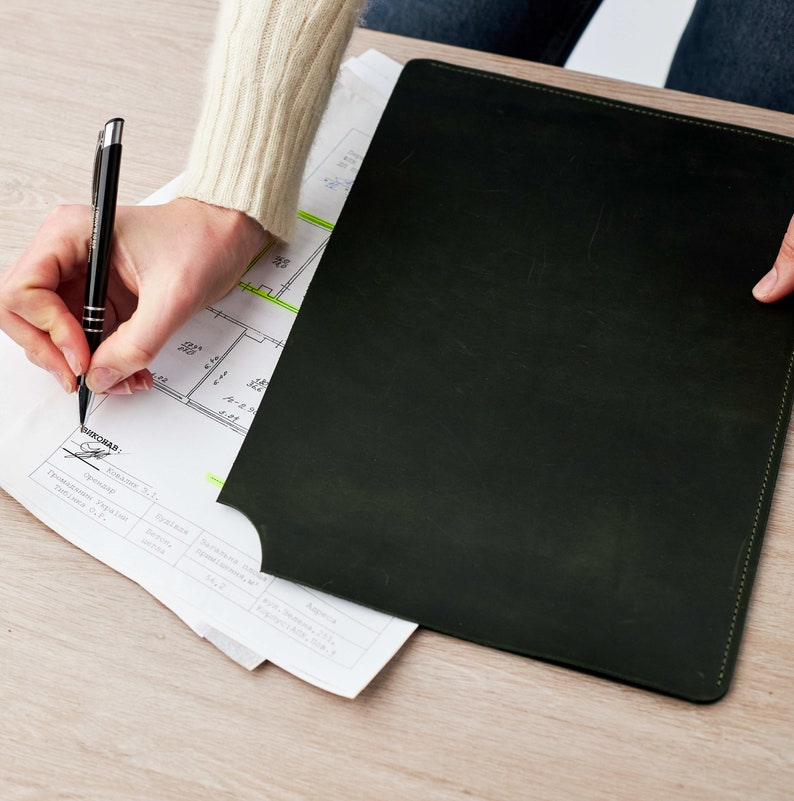 May include: A person signing a document with a black pen. The document is a blueprint with a green highlighter marking a section. The person is sitting at a desk with a black leather portfolio.