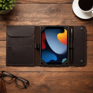 May include: Dark brown leather tablet case with a built-in stand, pen holder, and multiple pockets. The case is open, revealing a tablet with a colorful display. A black pen is in the holder. The case is on a wooden surface.