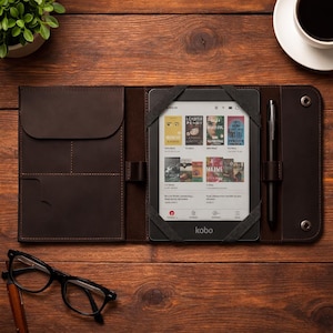 Pode incluir: Uma capa de couro castanho para leitor de livros eletrónicos com um leitor Kobo no interior, apresentando capas de livros no ecrã. Uma caneta preta está presa no lado direito. A capa está aberta numa superfície de madeira, com óculos e uma chávena de café por perto.