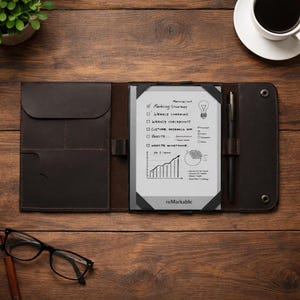 以下が含まれることがあります： 手書きのメモと図が表示されたデジタルタブレットが入った、濃い茶色の革製フォリオケース。黒いペンがタブレットの横に置かれています。タブレットの画面には「reMarkable」の文字が表示されています。フォリオは木製の表面で開いています。