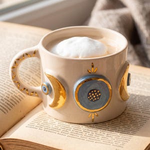 May include: A cream-colored ceramic mug filled with a frothy beverage, featuring gold-painted crescent moon and circular motifs, a small blue evil eye emblem, and gold leaf accents on the handle, resting on an open book with printed text.