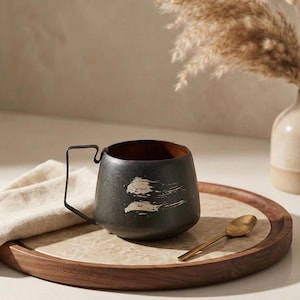 May include: A black ceramic mug with a distinctive handle and white brushstroke design, on a wooden tray with a marble effect. A small gold spoon is next to the mug. The mug's interior is dark brown.