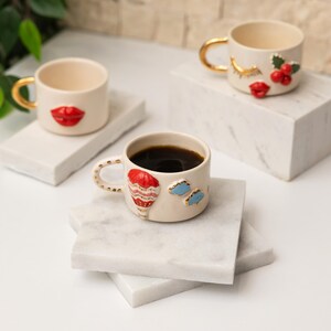 May include: Three white ceramic coffee mugs with gold handles and decorative details. One mug has a red lip design, another has a red and blue hot air balloon design, and the third has a red lip and red berries design.