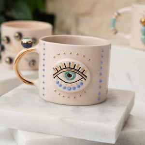 May include: A white ceramic mug with a gold rim and handle. The mug has a blue and green evil eye design with gold accents.