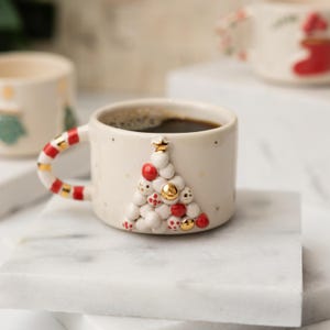 May include: White ceramic coffee mug with a red and white candy cane handle. The mug has a 3D Christmas tree design with red and gold ornaments.