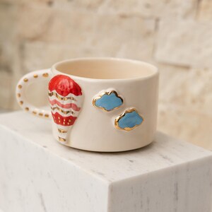 May include: A white ceramic mug with a red, white, and gold hot air balloon design and two blue clouds with gold accents.