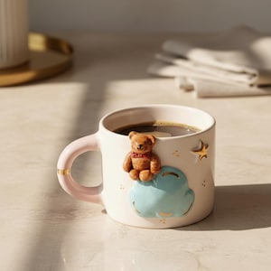 Pode incluir: Caneca de cerâmica branca cheia de café, com uma alça rosa claro e detalhes dourados. A caneca é decorada com um ursinho de pelúcia castanho, uma nuvem azul e uma estrela dourada. A caneca está sobre uma superfície clara.